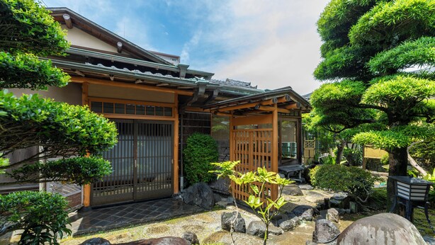 A traditional Japanese house with a tiled roof, wooden gate, and a garden with rocks and trees. - IKONASOU (Izunokuni)