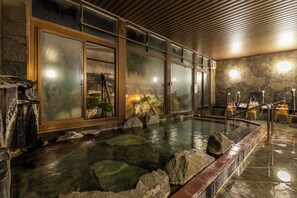 A traditional Japanese onsen with a stone interior, wooden benches, and a view of the outside through large glass doors. - IKONASOU (Izunokuni)