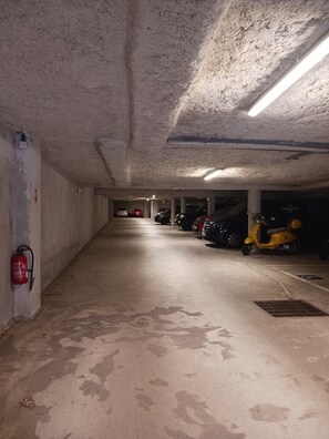 Un parking avec des murs en béton, un extincteur et plusieurs véhicules stationnés, dont un scooter jaune.