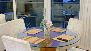 A round glass table with four white upholstered chairs, a vase with flowers, and a remote control on the table.
