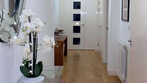 A hallway with a wooden floor, a glass-top table with a vase of white flowers, a mirror, and a door with a window.