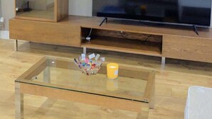 A living room with a large flat-screen TV mounted on a wooden wall unit. There is a glass coffee table in the center with a few items on it.