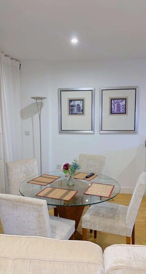 Un coin repas avec une table en verre entourée de chaises rembourrées, des tableaux encadrés au mur et un lampadaire.