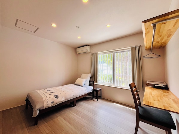 A small, single-bed room with a wooden desk and chair, a window with blinds, and an air conditioning unit. - Beach side Pension mibal (Nanjo)
