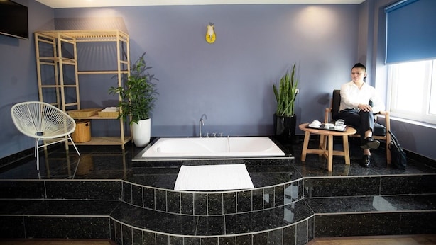 A spa area with a large bathtub, a wooden shelf with baskets, a white chair, and a potted plant. - Huong Duong Hotel Yen Xa by Bay Luxury (Hanoi)
