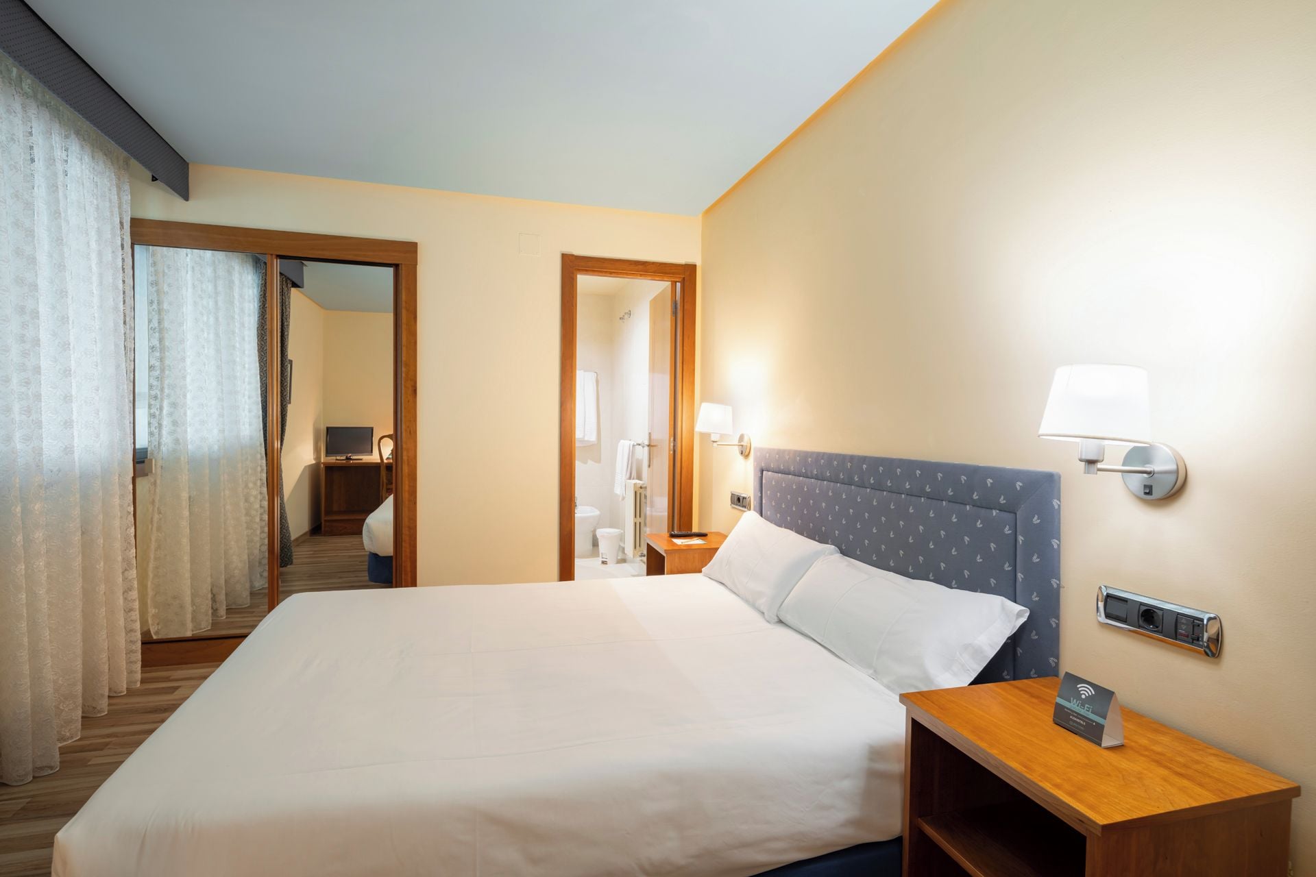 Habitación de hotel con una cama grande, una mesita de noche con lámpara, un espejo y vistas al baño.