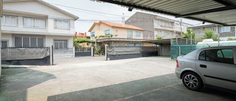 A covered parking area with a car parked on the right. There are houses in the background with visible balconies and windows.