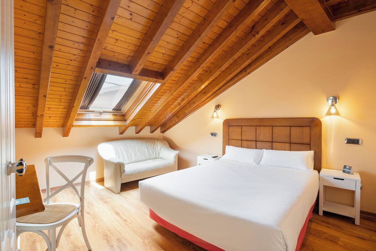 Un dormitorio con techo de madera, una cama, una silla y mesitas de noche.