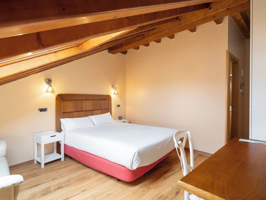 A bedroom with a wooden ceiling, a bed with white bedding, a nightstand, a bedside table, and a wooden desk.
