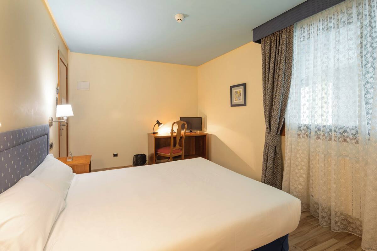 Una habitación de hotel con cama, escritorio, silla, lámpara y cortinas.