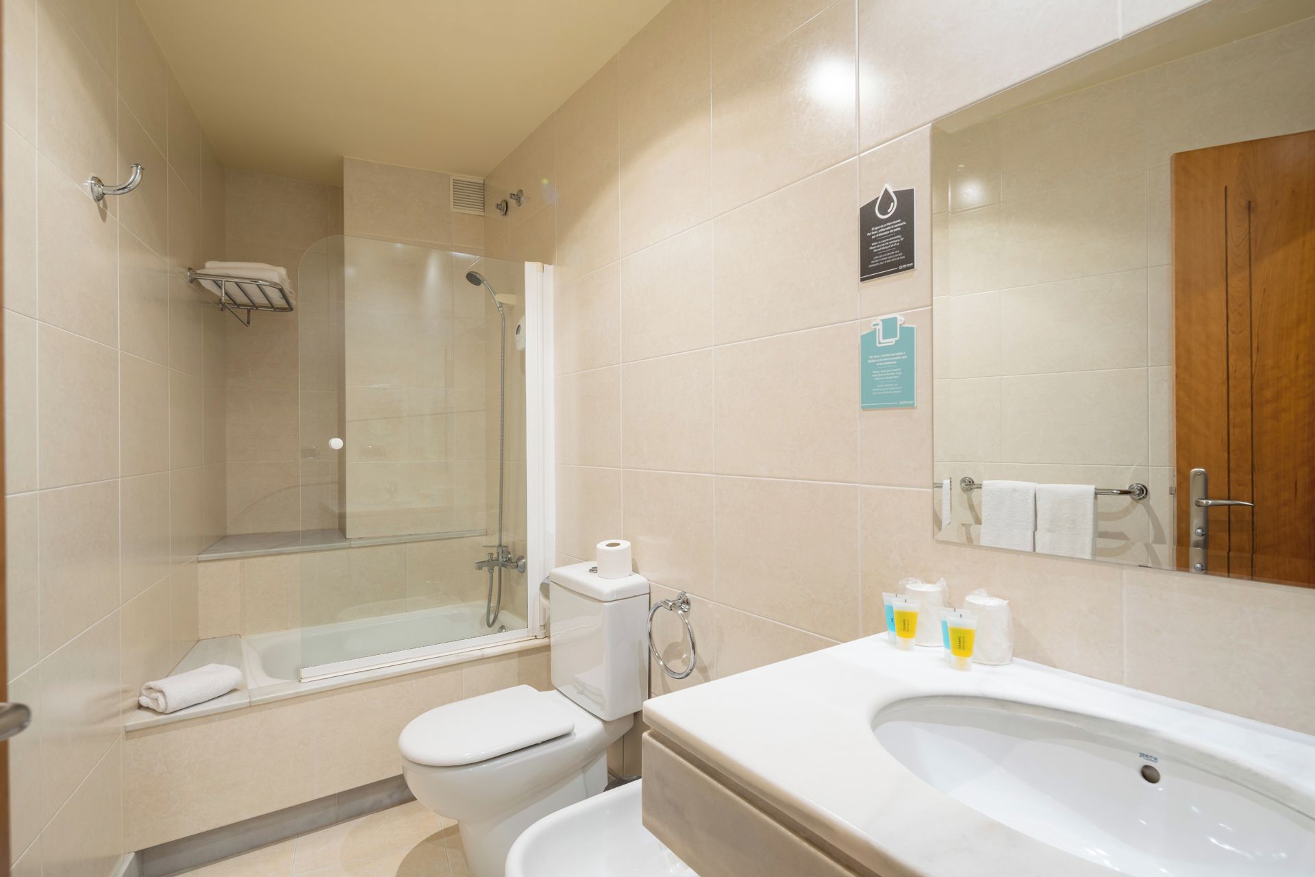 A modern bathroom with a glass-enclosed shower, a toilet, a sink with toiletries, and a wooden door.