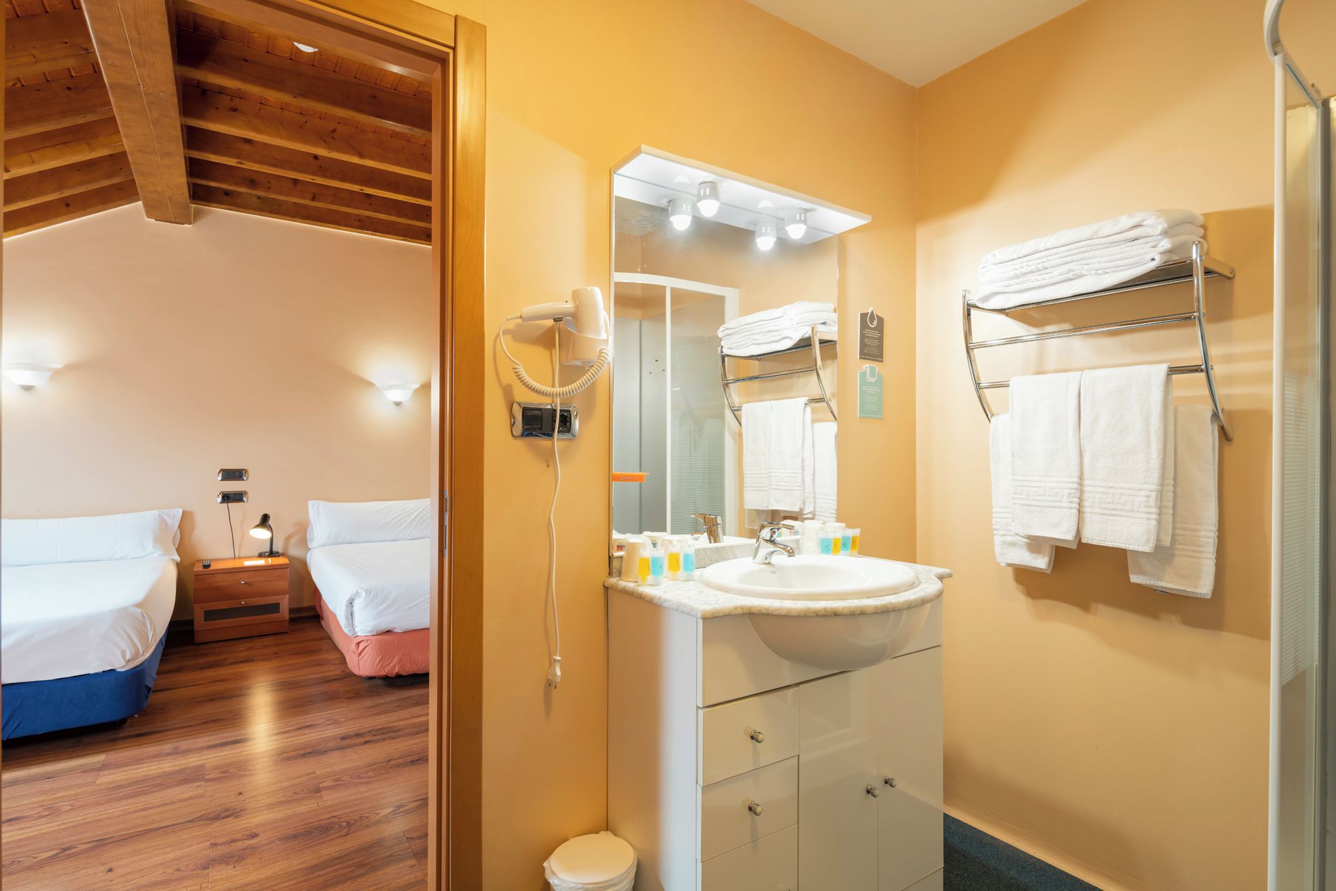Una habitación de hotel con techo de madera, una cama, y un baño con lavamanos, espejo y toallero.