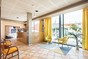 Vestíbulo de hotel con mostrador de recepción, sillas amarillas, un ventanal con cortinas amarillas y vistas a edificios.