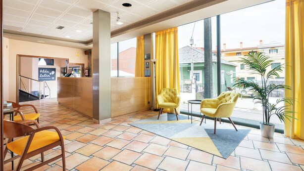 A hotel lobby with a reception desk, yellow chairs, a large window with yellow curtains, and a view of buildings outside. - Alda Lalín (Lalin)
