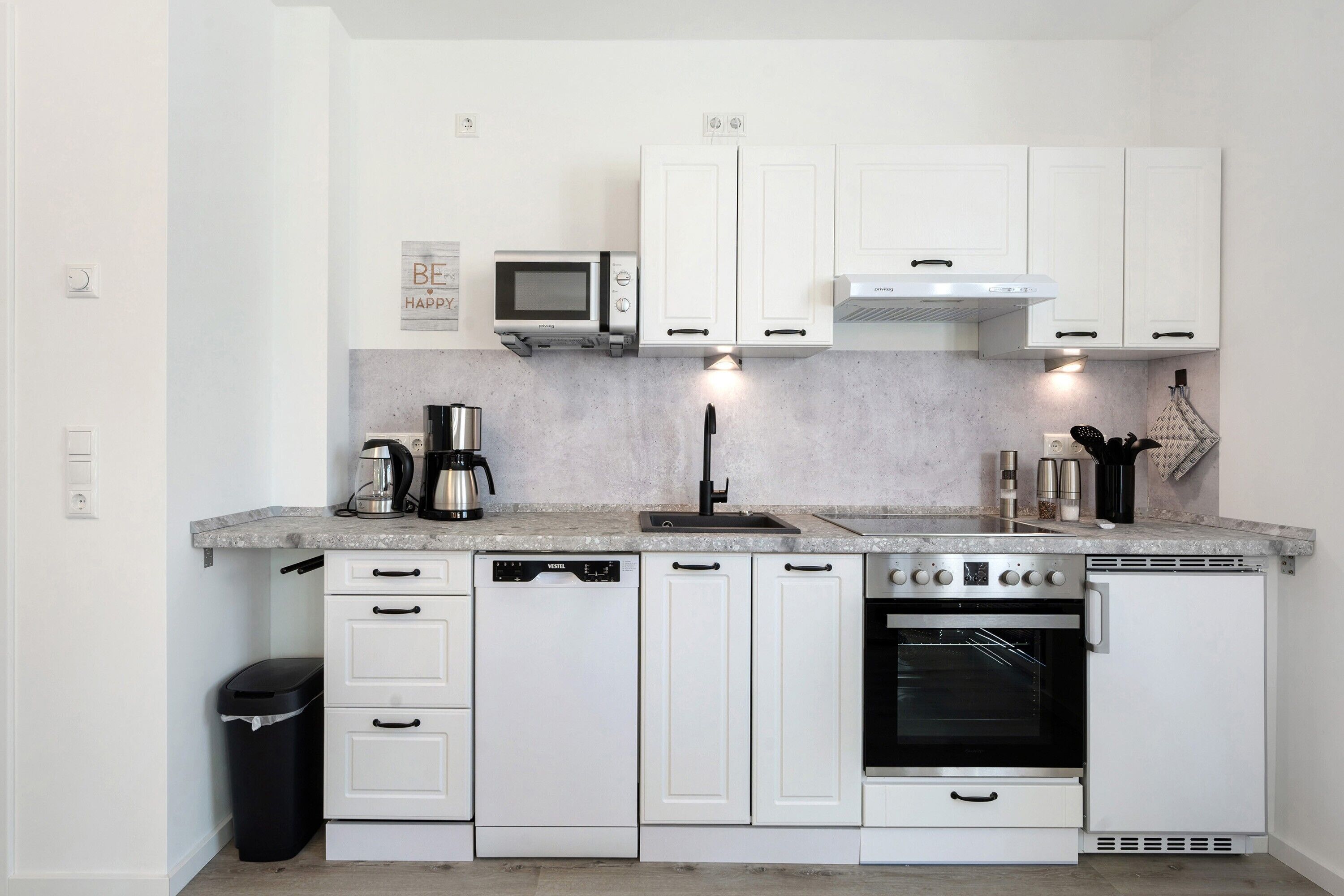 Cocina compacta con armarios blancos, microondas, cafetera, lavavajillas y horno.