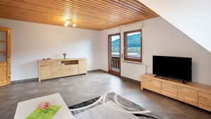 Une pièce spacieuse avec un plafond en bois, une grande fenêtre, un téléviseur et un meuble en bois.
