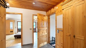 Un couloir en bois mène à une chambre à coucher où se trouvent un lit, une porte en bois ornée d’un élément décoratif et une petite table sur laquelle est posé un tapis.