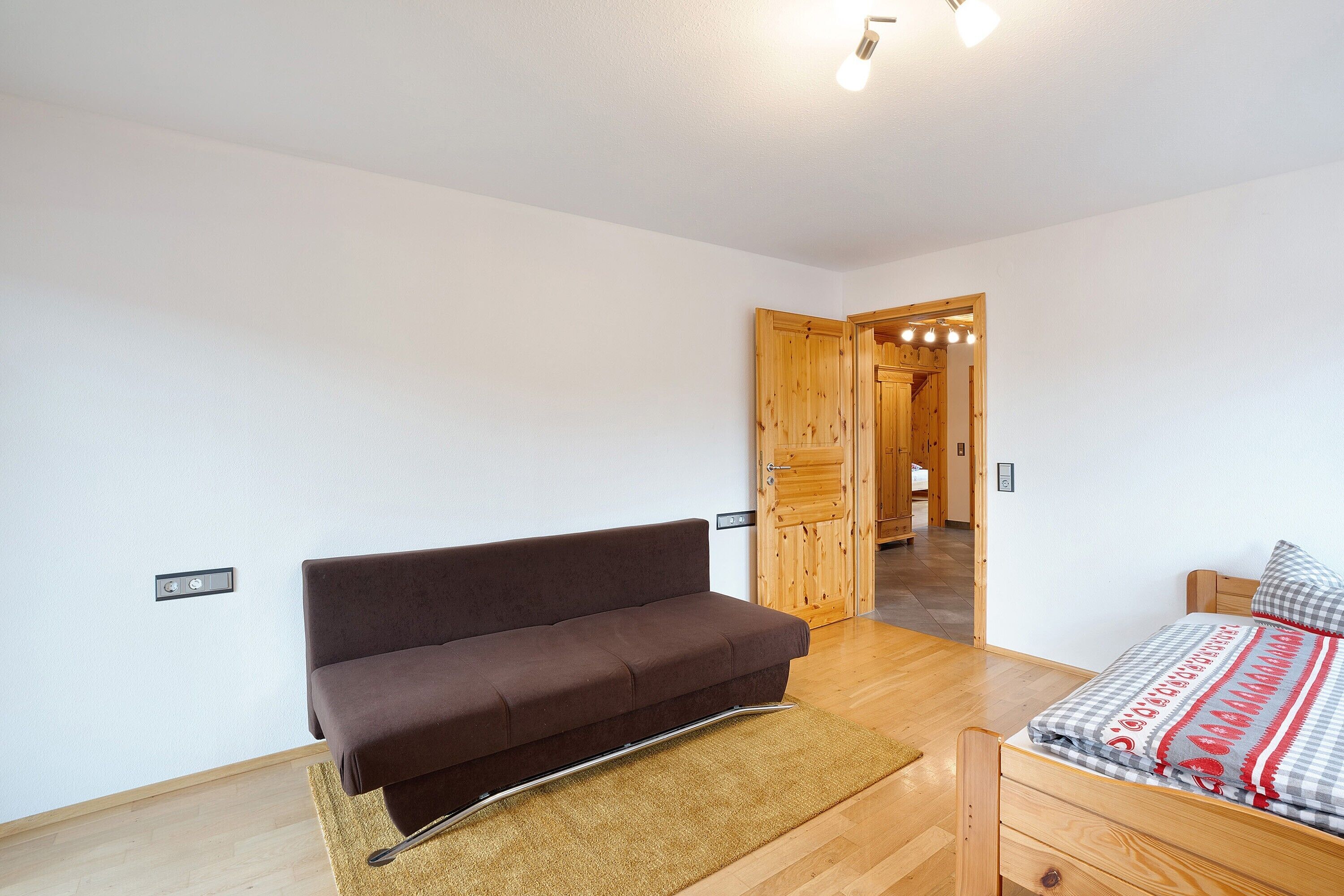 Una habitación con un sofá marrón, una cama con una manta estampada, una puerta de madera y un interruptor de luz en la pared.