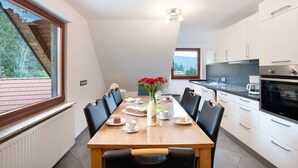 Un coin repas avec une table dressée pour six personnes, un vase rempli de fleurs rouges et une vue sur un bâtiment aux toits rouges à l’extérieur.