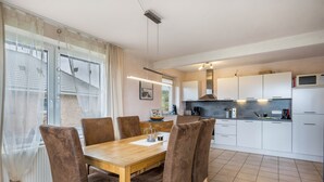 A dining area with a wooden table and chairs, a kitchen with white cabinets and appliances, and a view through a window with sheer curtains. - Sustainable Holiday Apartment "Lichtblick" with Private Terrace, Garden, Wi-Fi & EV Charger (Grömitz)