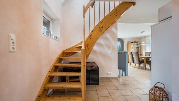 A wooden staircase in a room with tiled flooring, a window, and a dining area in the background. - Sustainable Holiday Apartment "Lichtblick" with Private Terrace, Garden, Wi-Fi & EV Charger (Grömitz)