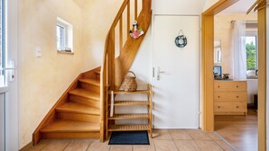 Un escalier en bois mène à une porte surmontée d’un panier et d’une étagère. Une fenêtre donne sur une pièce où l’on aperçoit une commode.