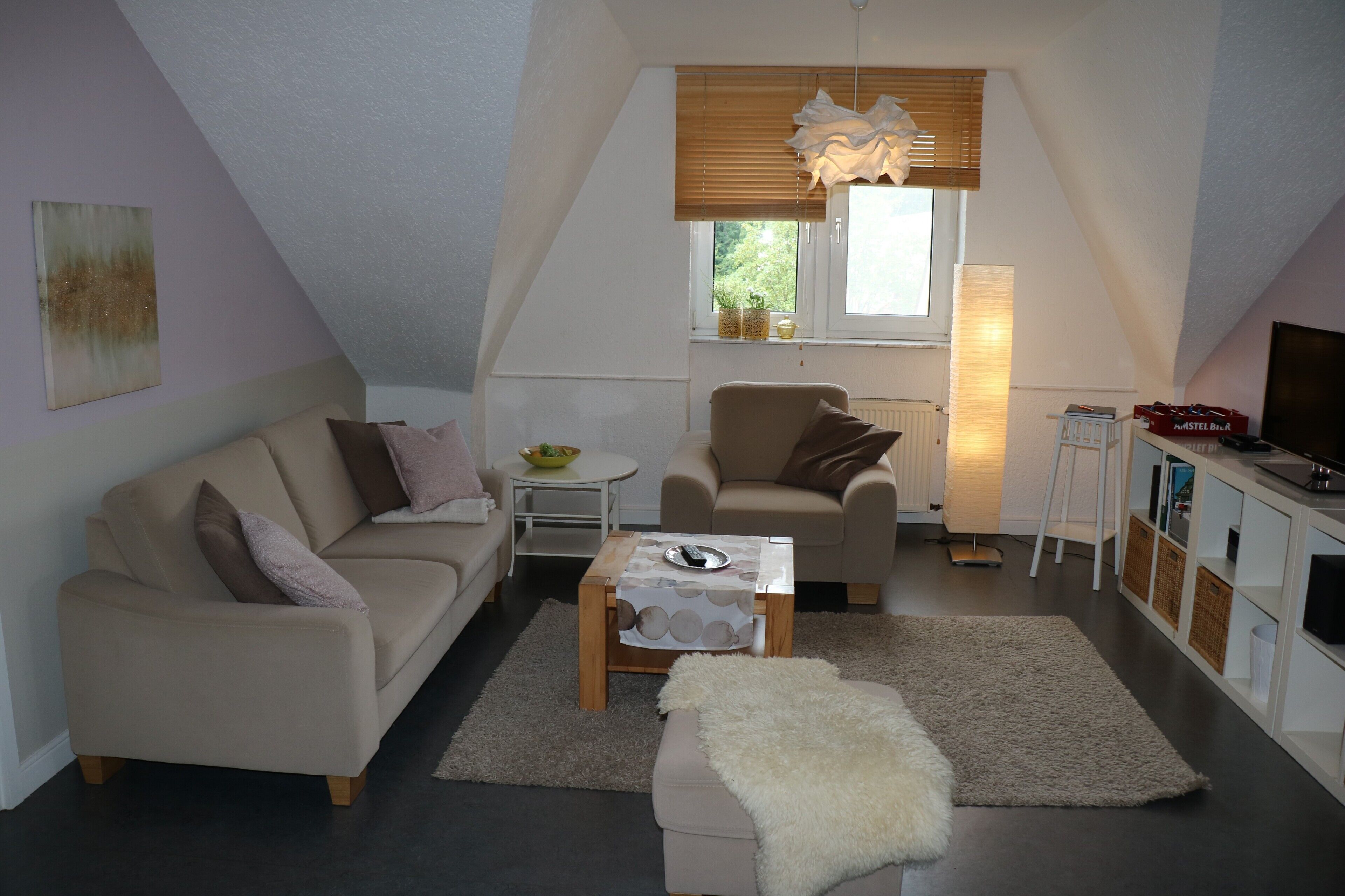 Ein Loft-Wohnzimmer mit hoher Decke, einer Couch, einem Sessel, einem kleinen Tisch, einem Fernseher und einem Fenster mit Jalousien.