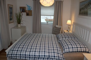 A bedroom with a bed, pillows, a nightstand, a lamp, a window with curtains, a radiator, and a wall-mounted shelf with a plant.