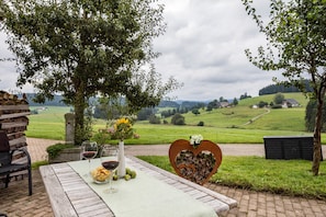 A rustic outdoor dining area with a table set for two, a heart-shaped wooden decoration, and a scenic view of a green landscape.