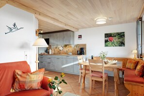 A cozy interior with a wooden ceiling, a dining area with a table and chairs, a kitchen with grey cabinets, and a red sofa with a plaid pillow.