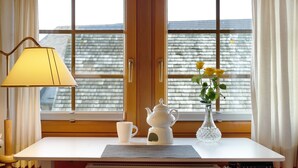 Ein Zimmer mit Tisch, Lampe, Teekanne, Blumenvase und einem Fenster mit Vorhängen.