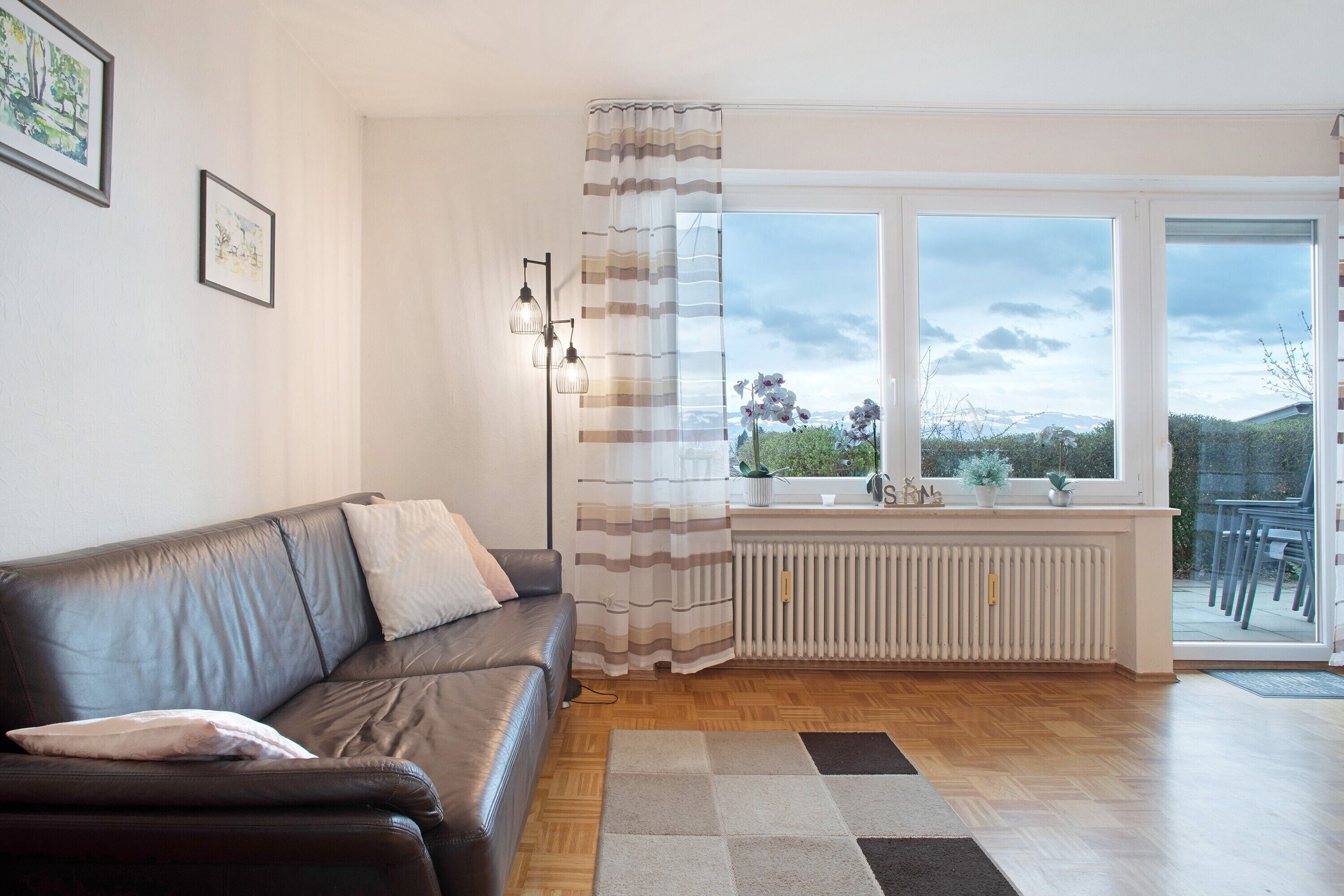 Ein Wohnzimmer mit einer Ledersofa, einem Heizkörper, einem Fenster mit Vorhängen und Blick ins Freie.