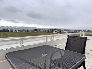 Un balcon avec une table et des chaises en métal, donnant sur un paysage rural avec des maisons et des champs.