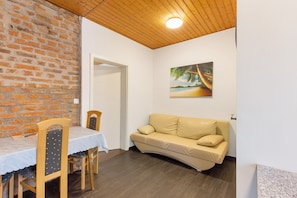 Een kamer met een bakstenen muur, een houten plafond, een witte deur, een beige bank, een eettafel met stoelen en een schilderij aan de wand.