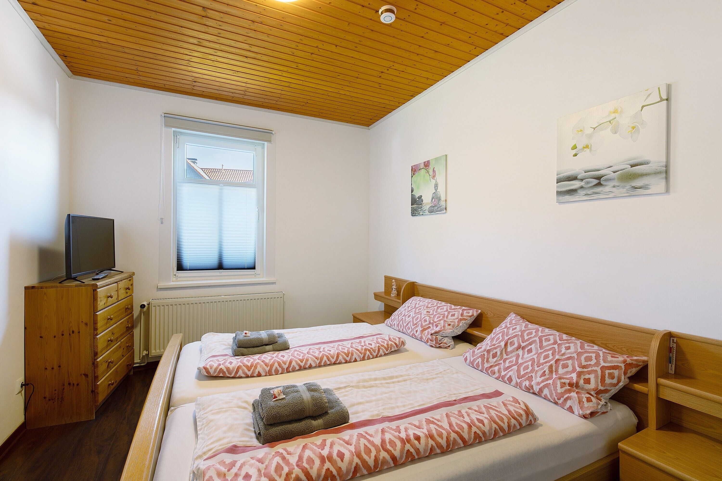 A bedroom with two beds, wooden headboards, a dresser, a television, and a window with blinds.