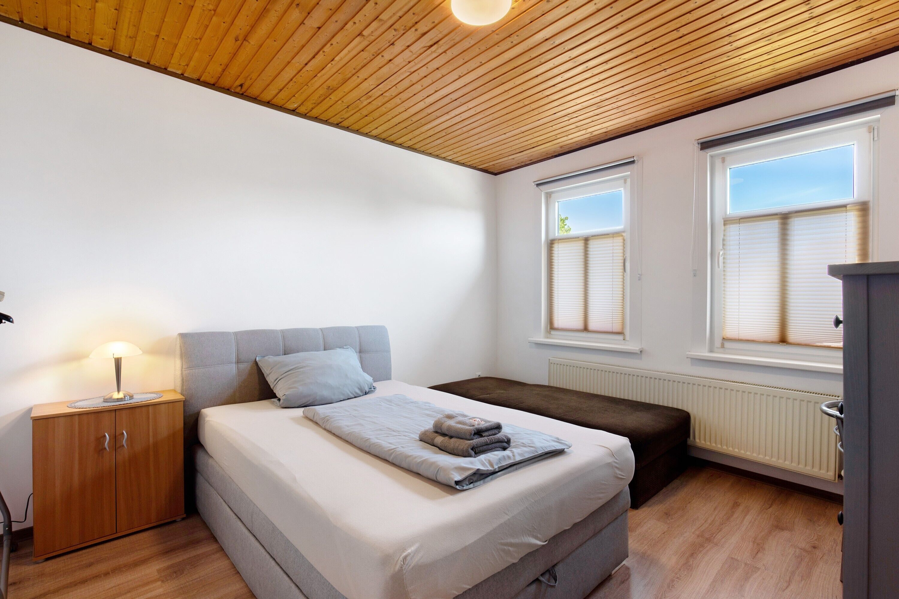A bedroom with a bed, bedside table, bench, and windows.