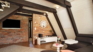 Un salón estilo loft con vigas de madera a la vista, pared de ladrillo, un reloj grande, un sofá de cuero negro y una mesa de centro redonda de vidrio.