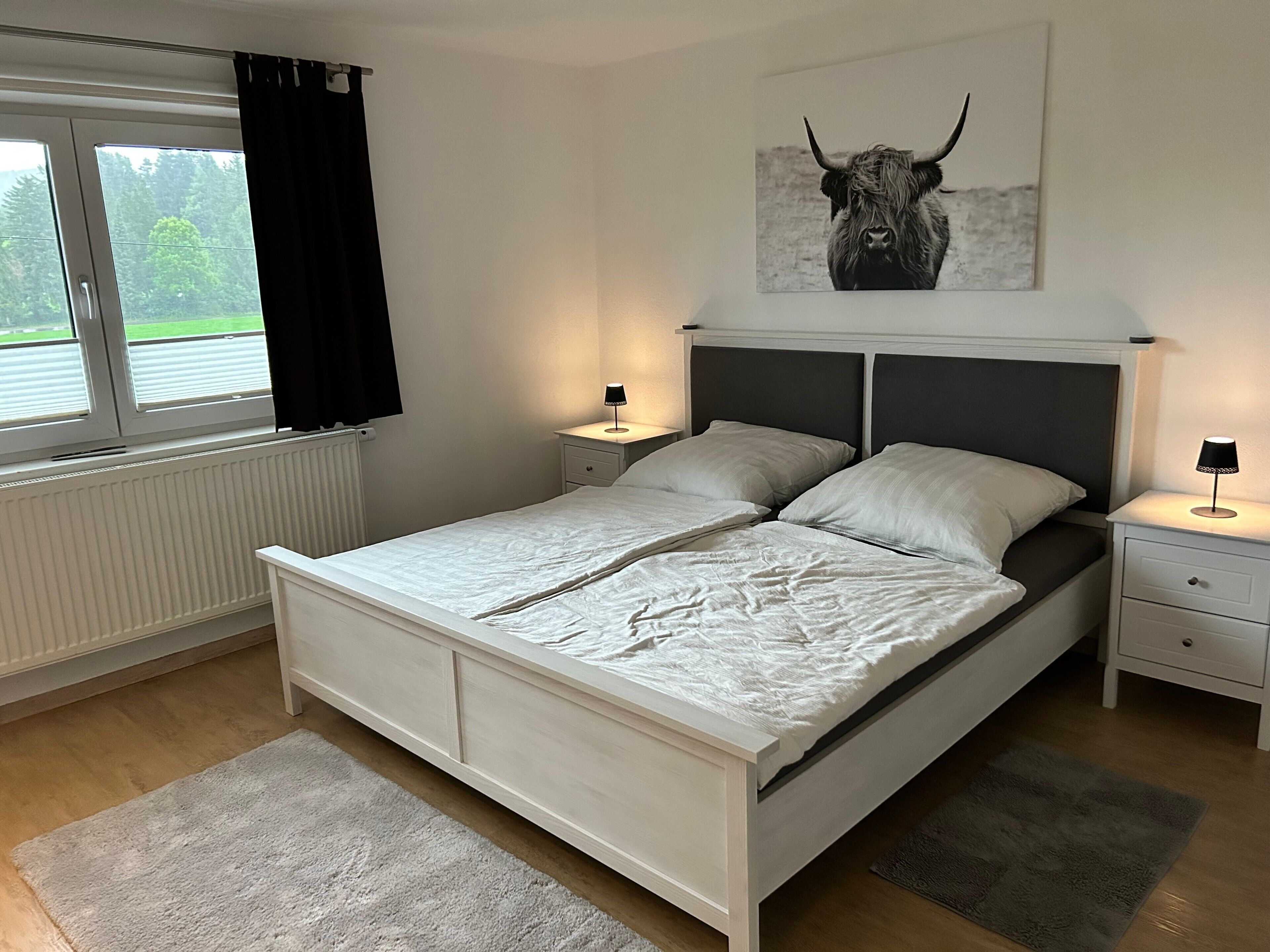 Une chambre à coucher comprenant un lit, deux tables de chevet, une fenêtre avec des rideaux, un radiateur et une œuvre d’art représentant un taureau accrochée au mur.