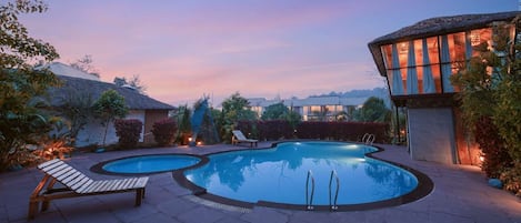 A pool area with two pools, lounge chairs, and a building with a thatched roof.