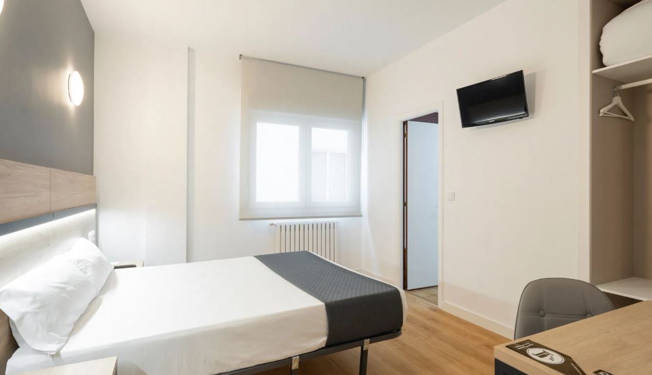 Habitación de hotel con cama, escritorio, silla, televisor y ventana con persianas.