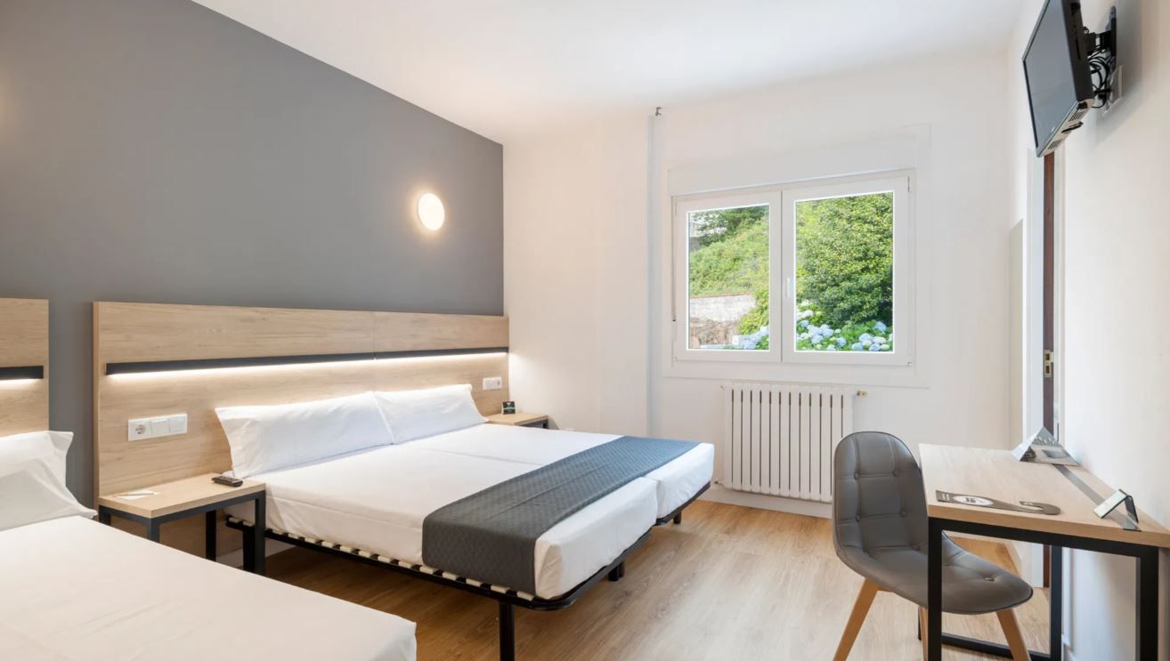Habitación de hotel moderna con una cama grande, mesitas de noche, un escritorio con silla y una ventana con vistas a zonas verdes.