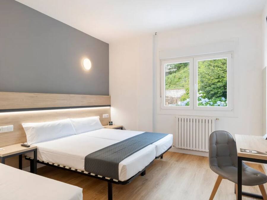 A modern hotel room with a large bed, bedside tables, a desk with a chair, and a window with a view of greenery.