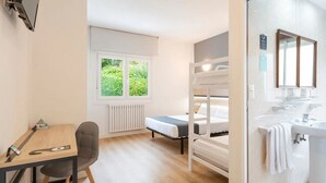 Una habitación con una cama individual doble, un escritorio con silla, una ventana con vistas a la vegetación y un baño con lavabo y espejo.