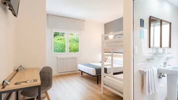 A room with a bunk bed, a desk with a chair, a window with a view of greenery, and a bathroom with a sink and mirror.