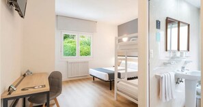 Una habitación con una cama individual doble, un escritorio con silla, una ventana con vistas a la vegetación y un baño con lavabo y espejo.