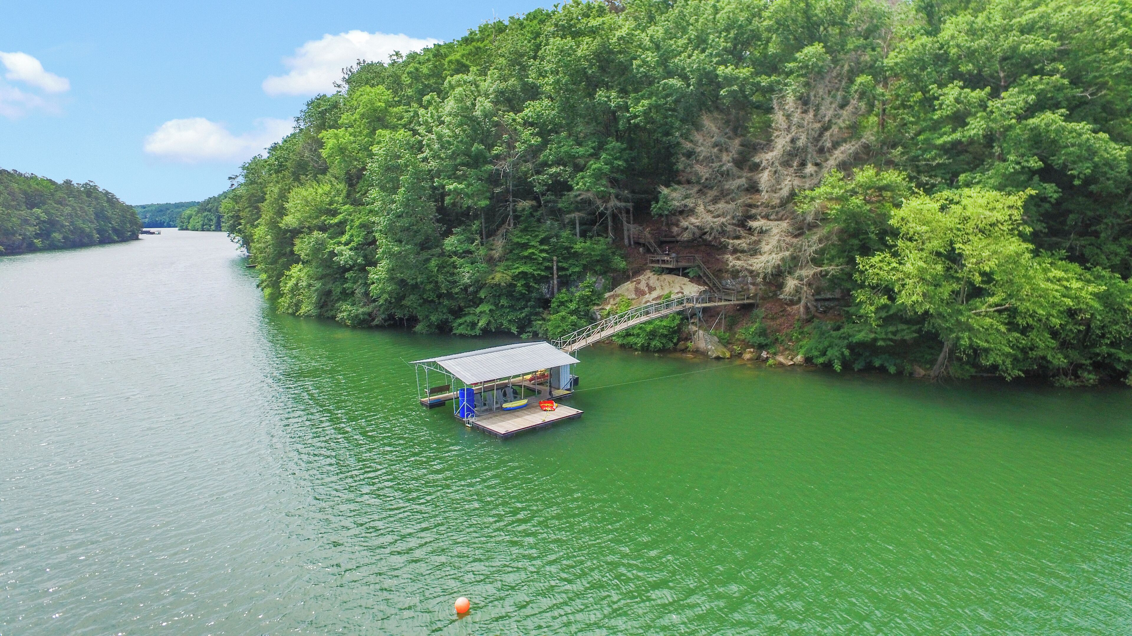 A docked boat with a covered seating area and a small building, surrounded by lush greenery and a steep cliff.