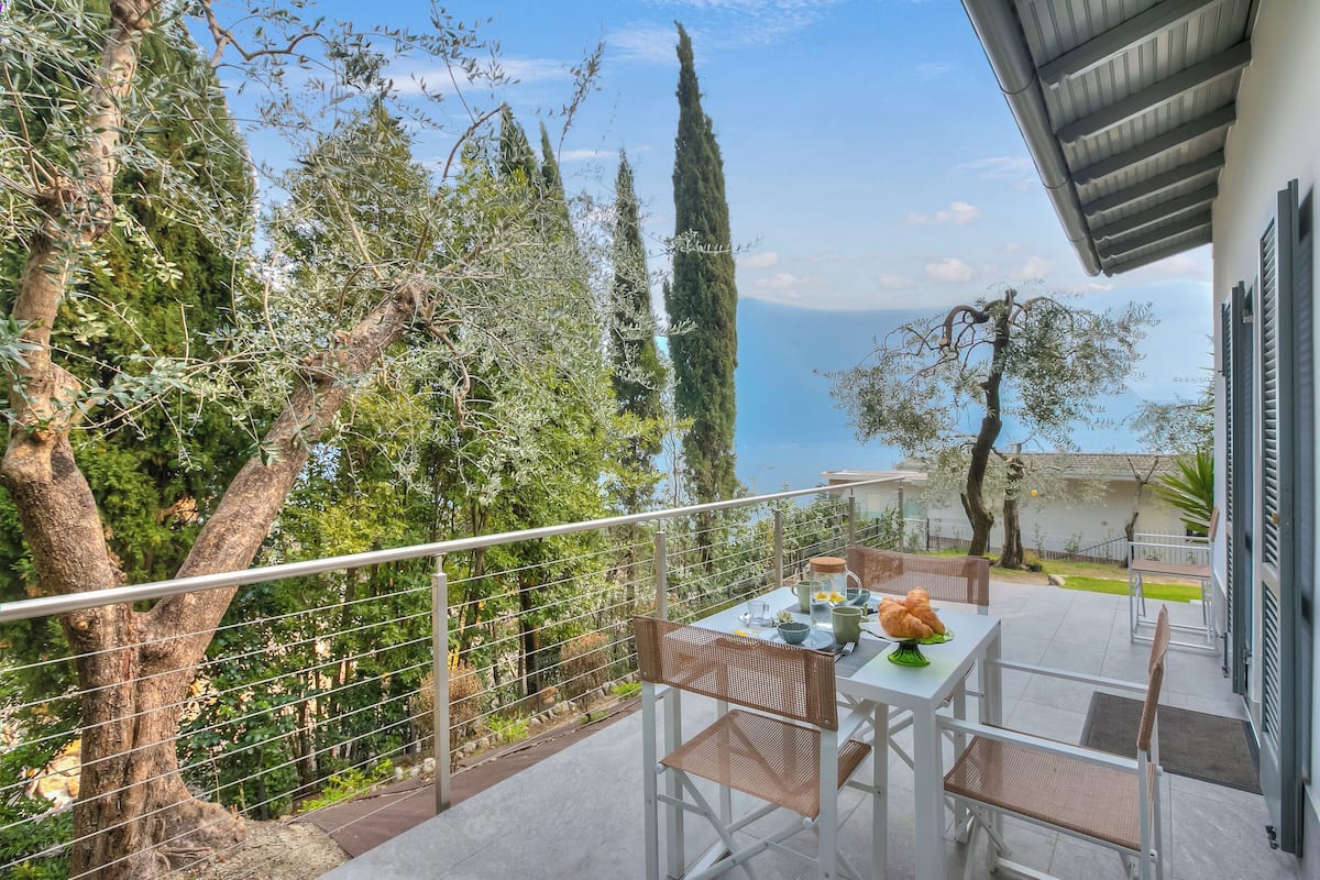 Un balcon avec une table dressée pour deux personnes, donnant sur un jardin et offrant une vue sur la mer.