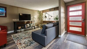 Un salon avec un canapé gris, un fauteuil rouge, un téléviseur posé sur un meuble noir, une porte rouge et une fenêtre avec des stores.