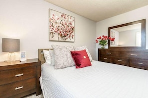 Quarto com cama, mesa de cabeceira, abajur, cômoda, espelho e pintura floral na parede.
