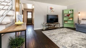 Ein Wohnzimmer mit dunklem Holzboden, einem grauen Teppich mit Muster, einer grünen Regaleinrichtung mit Dekorationen, einem blauen Sofa und einem Fernseher auf einem Schrank.